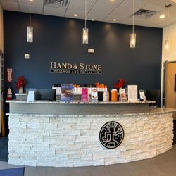 Hand & Stone Massage and Facial Spa - Elegant curved reception desk with stacked stone facade featuring Hand & Stone logo. Modern pendant lighting, branded wall si...