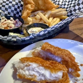 Effingbar And Grill - Fish & Chips - The image shows two fried fish fillets with a crispy golden-brown exterior and a creamy, white filling inside, placed on a white plate. In the background, there is a basket lined with checkered paper containing additional fish and chips, along with small containers of coleslaw and tartar sauce. The setting appears to be a casual dining environment, likely at a restaurant or pub specializing in seafood and bar food. - American Near Me - Albuquerque, New Mexico