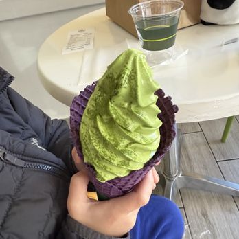 Matcha Café Maiko - Matcha 01. Soft-Serve Ice Cream - A person, wearing a gray jacket and blue pants, holds a large swirl of green matcha soft serve ice cream in a purple waffle cone. The background shows a white table with a cup of dark green beverage, a receipt, and a paper bag, indicating a cafe setting. The ice cream has a smooth, swirled texture and is topped with matcha powder. - Bubble Tea Near Me - Boston, Massachusetts