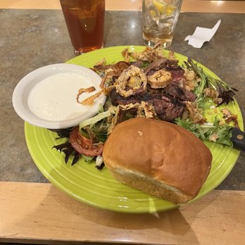 O'Niell's - The image shows a meal served on a bright green plate, featuring a sandwich bun, a mixed salad with tomatoes and greens, and fried onion rings. A small dish of creamy dressing is placed on the plate. Behind the plate, there is a glass of iced tea with lemon and a glass of water with lemon slices. The setting appears to be a casual dining environment with wooden chairs and a table surface. - Irish Near Me - Albuquerque, New Mexico