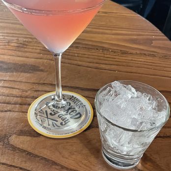 The Tap - Cosmo! - A pink cocktail garnished with a lime wedge is placed on a wooden table, alongside a glass of ice water. - Pubs Near Me - Cumberland, Rhode Island