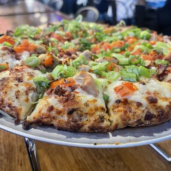 Mountain Mike's Pizza - A close-up of a pizza topped with melted cheese, chopped green onions, and various vegetables, served on a metal stand. The background shows a busy restaurant interior with patrons seated at tables, some wearing casual clothing. The pizza appears freshly baked with a crispy crust and colorful toppings, and the setting suggests a casual dining environment. - Pizza Near Me - Orem, Utah