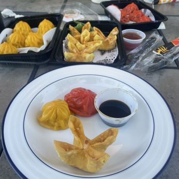 Umami - Take out - The image shows a table with multiple dim sum dishes, including yellow steamed dumplings, a red steamed bun, and a fried dumpling with dipping soy sauce. Additional takeout containers with more dumplings and sauces are visible in the background, along with a white cup and utensils. The setting appears to be an outdoor or casual dining area. - Ramen Near Me - Annapolis, Maryland