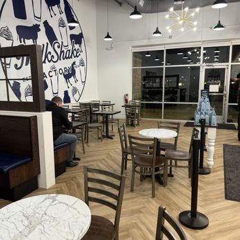MilkShake Factory Arvada - Front of House - The image shows the interior of a milkshake shop with wooden flooring, several tables and chairs arranged throughout the space, and a large circular logo on the wall displaying the shop's name and images of milkshakes and cows. The ceiling has exposed ductwork and hanging lights, with a modern chandelier near the entrance. Large glass doors lead outside, and a person is seated at one of the tables. The shop has a clean, contemporary design. - Ice Cream & Frozen Yogurt Near Me - Arvada, Colorado