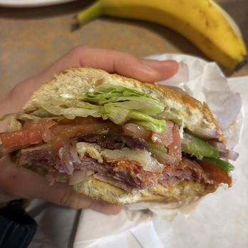 Zing! Cafe - Italian - A hand is holding a sandwich with visible layers of lettuce, tomato, and meat inside a bun. A banana is placed on the table behind the sandwich, and part of a white plate with food is also visible in the background. The setting appears to be a casual dining area or kitchen. - Coffee & Tea Near Me - Berkeley, California