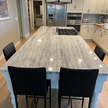 Davan Countertops - A modern kitchen features a large granite island with four black chairs, white cabinetry, and stainless steel appliances including a refrigerator and oven. - Kitchen & Bath Near Me - Franklin Park, Illinois