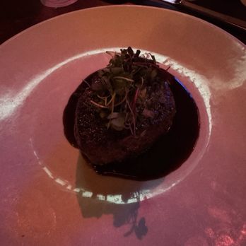 The Precinct - Herb Crusted Filet - A cooked steak garnished with microgreens is presented on a white plate with a dark sauce underneath, in a dimly lit restaurant setting. - Steakhouses Near Me - Cincinnati, Ohio