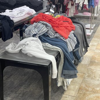 Macy's - A retail clothing store displays various garments on tables and racks. Folded shirts and pants are arranged on a black table, while other clothing items, such as dresses and blouses, hang on racks in the background. The store has a tiled floor and a mix of casual and formal apparel, with some items appearing disorganized or in the process of being sorted. - Department Stores Near Me - Alpharetta, Georgia