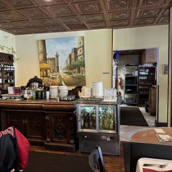 Pasquale Rigoletto Restaurant - The photo depicts the interior of a restaurant featuring a wooden bar with various bottles, glasses, and plates. A large painting of a cityscape hangs on the wall behind the bar. To the right, there is a doorway leading to a kitchen or storage area, with shelves and equipment visible. The ceiling has decorative tin tiles, and part of a table with utensils is visible in the foreground. - Italian Near Me - Bronx, New York