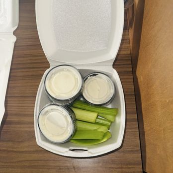 Union Jack's Pub - 8/15/25 - Celery & Ranch Dressing - A foam food container is open, revealing celery sticks and three small cups filled with ranch dressing. The container is situated on a wooden table, with part of a white foam container visible on the left side. The background includes some objects and a brown paper surface on the right side. - Pubs Near Me - Glenside, Pennsylvania