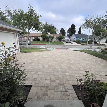 Restore & Construct - A paved residential driveway extends from the house to the street, bordered by garden beds with plants and flowers, with trees and houses visible across the street. - Roofing Near Me - Carson, California