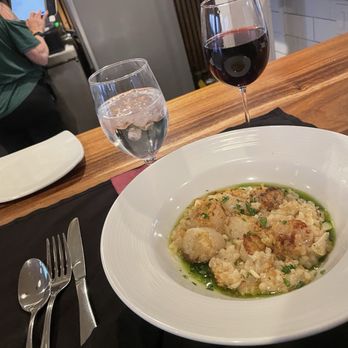 Cellar 417 - Scallops with basil oil - The image shows a restaurant table with a white plate of risotto containing scallops and garnished with herbs, placed on a black napkin. There is a glass of red wine and a glass of water with ice on the table. In the background, a person wearing a green shirt is using a touchscreen device, and part of the restaurant interior with wooden accents is visible. - Wine Bars Near Me - Branson West, Missouri