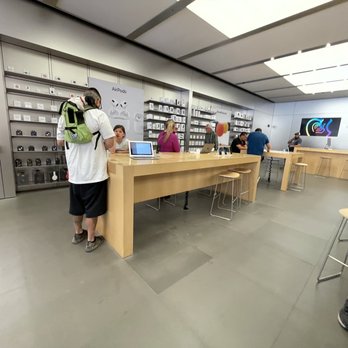 Apple ABQ Uptown - The image shows the interior of an Apple Store, featuring a long wooden counter where customers are being assisted by staff. The wall behind the counter displays various Apple products, including AirPods, with a large digital screen on the right side. Several customers are seated or standing, engaging with staff or browsing products. The store has a modern, minimalist design with bright lighting and a clean, organized layout. - Computers Near Me - Albuquerque, New Mexico