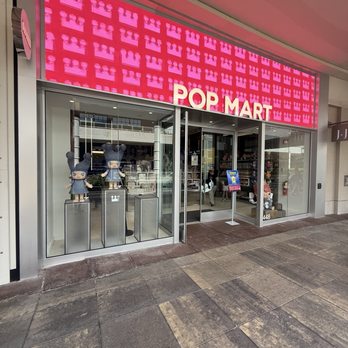 Pop Mart - The image shows the exterior of Pop Mart, a retail store, with a large pink digital sign displaying a repeating pattern and the store name in bold letters. The storefront has a glass entrance with display windows showcasing collectible figures on pedestals. The sidewalk in front is paved with large stone tiles, and the interior of the store is partially visible through the glass. - Toy Stores Near Me - Oak Brook, Illinois