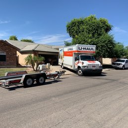 Backed in and ready for the movers! - A U-Haul moving truck and trailer are parked on a suburban street in front of a single-story house with a brick and stucco exterior. The truck is white with orange and black branding, and the trailer is unhitched and positioned on the road. There are several other vehicles parked along the street, and a large tree provides shade near the house. The sky is clear and blue.
