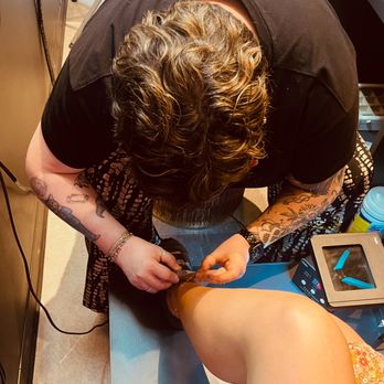 Peppermint - A person with tattoos on their arms is seated with one leg raised, receiving a pedicure from a technician. The technician is focused on the person's toenails, using tools and a device on a small table. The setting appears to be a salon, with various supplies visible nearby. - Women's Clothing Near Me - Rochester, New York