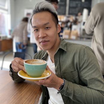 Haven Brews - Aesthetics - A young man with styled, two-tone hair (dark at the roots and silver at the tips) is seated at a wooden table in a cafe, holding a ceramic coffee cup and matching saucer. He wears a green jacket over a white textured shirt and has a watch on his left wrist. The background shows other patrons, a menu board, and cafe interior details, with a shallow depth of field focusing on the individual. - Coffee Roasteries Near Me - Toronto, Ontario