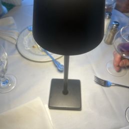 Light - A black lamp shaped like a wine glass is positioned on a dining table covered with a white tablecloth. The table has various items including wine glasses, a plate with food remnants, utensils, and salt and pepper shakers. The setting appears to be at a restaurant or a formal dining event, with other people partially visible in the background.