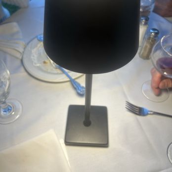 Casa Bella - Light - A black lamp shaped like a wine glass is positioned on a dining table covered with a white tablecloth. The table has various items including wine glasses, a plate with food remnants, utensils, and salt and pepper shakers. The setting appears to be at a restaurant or a formal dining event, with other people partially visible in the background. - Italian Near Me - Scranton, Pennsylvania