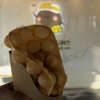 TP Tea - Original Egg Puffs - A bubble waffle with white chocolate chips is held in front of a wall featuring a cartoon character illustration, likely at a dessert shop. - Bubble Tea Near Me - Millbrae, California