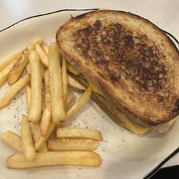 Open Tuna Melt - A grilled cheese sandwich with toasted bread and melted cheese is placed on a white plate. The sandwich appears to be cut in half, with the melted cheese visible at the edges. French fries are served alongside the sandwich, seasoned with black pepper. The plate has a black rim and is set on a light-colored table surface.