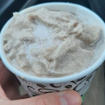Cafe Cubano - Banana and Peanut Butter Frappe - A hand is holding a paper cup filled with coffee that has foam and liquid on top. The background shows part of a vehicle's interior, including the driver's side door with window controls and a steering wheel. The image captures a casual moment of enjoying coffee inside a car. - Cuban Near Me - Springfield, Missouri