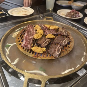 Boon Dee Moo Ka Ta - A round hotpot with grilling meat, seafood, and vegetables is on a table with diners, plates, and drinks. - Thai Near Me - Flushing, New York