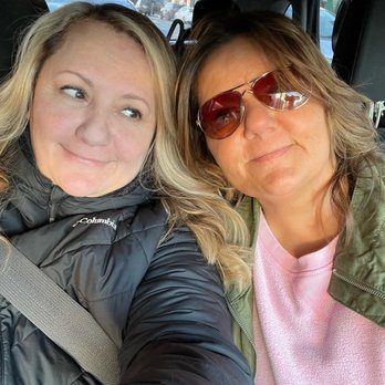Epoch Studio Salon - Chicago - Two women are seated in the back of a car, taking a selfie. One woman has blonde hair, is wearing a black Columbia jacket, and is smiling while looking at the other woman. The second woman has brown hair, is wearing sunglasses, a pink shirt, and a green jacket, and is smiling slightly. The background shows car seats and windows with a view of parked cars outside. - Hair Extensions Near Me - Chicago, Illinois