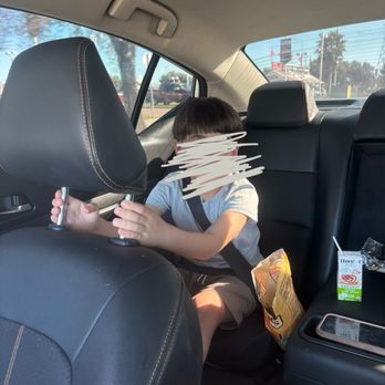 Clipper King - A young boy is seated in the back row of a car, wearing a seatbelt, and holding onto the headrest of the front passenger seat. The car interior features black leather seats, and there are snacks and a smartphone placed on the center console. The background outside the car shows a sunny day with trees and buildings visible through the windows. - Barbers Near Me - Tracy, California