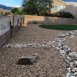 a gravel path with a rock in the middle - A backyard features gravel ground, a rock pathway, and a small artificial grass area, enclosed by a brick and metal fence.