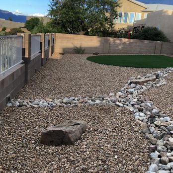 AMG Landscape - a gravel path with a rock in the middle - A backyard features gravel ground, a rock pathway, and a small artificial grass area, enclosed by a brick and metal fence. - Irrigation Near Me