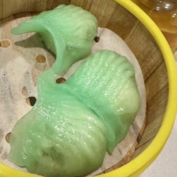Ming Mun - Two green rice rolls shaped like dolphins are arranged in a bamboo steamer basket lined with parchment paper. The rice rolls have a shiny surface and detailed dolphin features, including fins and a curved tail. The basket has a yellow rim and is set on a table with some bottles visible in the background. The image highlights the presentation of dim sum items in a traditional bamboo steamer. - Dim Sum Near Me - Brooklyn, New York