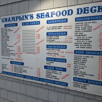 Champlin's Seafood - Menu - A large outdoor menu board for Champlin's Seafood in Narragansett, RI, displays various seafood dishes, sides, drinks, and draft beers. The menu includes items like fried clams, lobster rolls, steamed clams, and raw bar options, with prices and notes on daily specials. Beverage options include coffee, soda, bottled water, and draft beers such as domestic draft, Corona Light, and craft beers. The board is mounted on a gray shingled wall and features blue and red text for different sections and menu items. - Seafood Near Me - Narragansett, Rhode Island