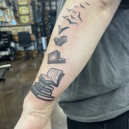 The tattoo features a stack of books at the bottom, with open pages, transitioning into a series of birds flying upward and outward. The design is in black ink with fine line work, situated on the inner forearm of a person wearing a dark shirt. The background shows a tattoo studio with shelves and chairs.