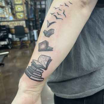 Double Down Tattoo - The tattoo features a stack of books at the bottom, with open pages, transitioning into a series of birds flying upward and outward. The design is in black ink with fine line work, situated on the inner forearm of a person wearing a dark shirt. The background shows a tattoo studio with shelves and chairs. - Tattoo Near Me - Glendale, Arizona