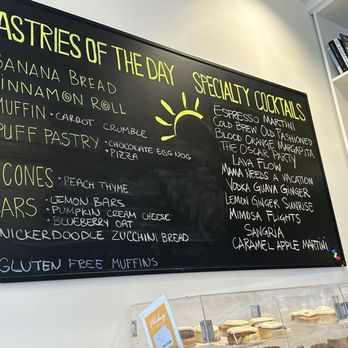 Perk Up Kitchen - Pastries and cocktails - A blackboard menu features sections for pastries and specialty cocktails, listing items such as banana bread, cinnamon roll, muffin, puff pastry, scones, bars, and various cocktails like espresso martini and blood orange margarita. The menu is written in colorful chalk and includes a small sun illustration. The setting appears to be a bakery or cafe with a display case of baked goods below and a shelf with plants and books on the right. - Breakfast & Brunch Near Me - Bloomington, Minnesota