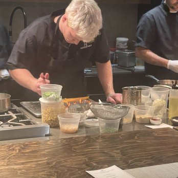 Hudson Table - Hoboken - David preparing the meal. - Two chefs are engaged in food preparation in a commercial kitchen setting. The chef on the left has blond hair and is leaning over the counter, focusing on their work, while the chef on the right, wearing glasses, is also involved in cooking. The counter is filled with containers of ingredients, utensils, and kitchen equipment. A menu is placed on the table in the foreground, and shelves with kitchen supplies are visible in the background. - Cooking Schools Near Me - Hoboken, New Jersey