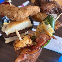 bacon, cheeseburger, and fried chicken skewers - A variety of grilled meats and vegetables on skewers are arranged in a tall glass, with some skewers resting on the rim, set on a dark wooden table.