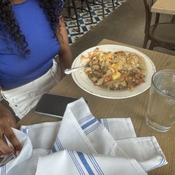 The Harvest Room - Mount Biscuit Skillet - A woman wearing a blue top and white pants is seated at a restaurant table with a plate of food and a glass of water. - New American Near Me - Munster, Indiana