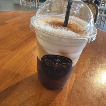 Queen B - Freddo Espresso - An iced coffee beverage with whipped cream, cocoa powder topping, and a black straw in a clear plastic cup, on a wooden table in a cafe. - Cafes Near Me - Chicago, Illinois