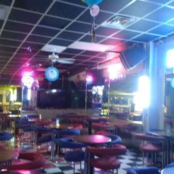 Silva Lanes-Eucan Bowl - The entrance of the bar. - The image shows the interior of a bar or nightclub with multiple tables and chairs arranged throughout the space. The ceiling has a grid pattern with hanging paper lanterns and decorations. Neon and colored lights illuminate the area, creating a vibrant atmosphere. There are ceiling fans and a large speaker mounted on the wall, with a checkered floor visible. The overall setting suggests a lively entertainment venue. - Bowling Near Me - Albuquerque, New Mexico