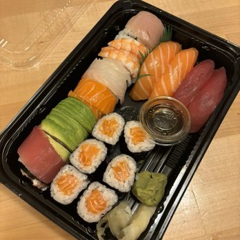 Manpuku - Sushi combo - A sushi platter in a black plastic container includes assorted sushi rolls with salmon, tuna, shrimp, and avocado, along with slices of sashimi such as salmon and tuna. The platter also contains sliced avocado, a small container of soy sauce, wasabi, and pickled ginger. The container is placed on a wooden surface, with a clear plastic lid partially visible in the top left corner. - Japanese Near Me - Berkeley, California
