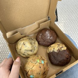 Cookies - A person holds a small cardboard box with four cookies inside. The cookies include two chocolate chip cookies, one double chocolate cookie with a dusting of powdered sugar, and one cookie with colorful candy pieces. The box is open, revealing the cookies, and the background shows a textured surface, possibly a table or countertop. The person's hand and part of their lap are visible, with the person wearing jeans.