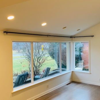 Goldak Construction - The image shows a living room with a large, multi-pane window providing a view of a garden with trees and outdoor seating. The room has hardwood flooring, white walls, and a ceiling with recessed lighting. Black curtain rods are installed above the windows, and there are vents along the baseboard. The outdoor area visible through the windows includes chairs and a brick wall. - General Contractors Near Me
