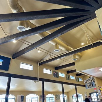 Sangio's Deli - Dining Room & Tasting Room Interior. - The image shows the interior of a restaurant with a high ceiling supported by dark wooden beams. The ceiling has exposed ductwork and small, rectangular windows near the top. Large glass doors and windows allow natural light to fill the space, revealing tables and chairs arranged for dining. Artwork is displayed on the walls, and a person is seen working at the counter area. The overall setting suggests a casual dining environment with a spacious and open layout. - Delis Near Me - Temecula, California