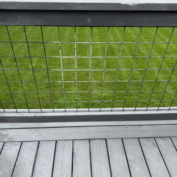 Performance Stoneworks - Mess from cutting stone on the new deck. - The image shows a wooden deck with gray planks and a metal wire fence railing, overlooking a green grassy field with trees in the background. - Countertop Installation Near Me - Indianapolis, Indiana