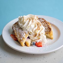 Stuffed French Toast - A plate features stuffed French toast slices topped with a swirl of whipped cream, dusted with powdered sugar. Fresh strawberries and blueberries are placed around the toast, and the dish is presented on a white, oval-shaped plate with a light-colored surface background.