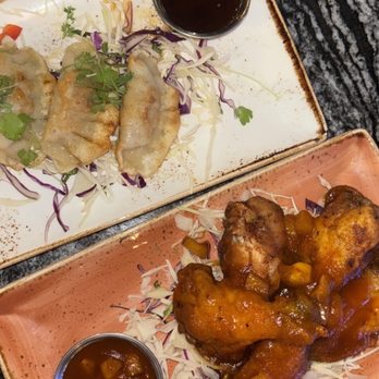 Ostras - The image shows two rectangular ceramic plates on a dark granite table. One plate contains three dumplings garnished with microgreens and shredded cabbage, with a small cup of soy sauce. The other plate has several chicken wings coated in a sauce, served with a small cup of dipping sauce. The dishes are presented on shredded cabbage and purple cabbage, highlighting Asian fusion cuisine. - Asian Fusion Near Me - Des Plaines, Illinois