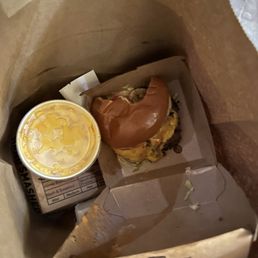 This is what we saw upon opening the bag.. - Inside a paper bag, there is a breakfast sandwich featuring a biscuit, egg, cheese, and sausage patty, with a bite taken out of the croissant on top. A small container of butter is also present. The items are arranged on a piece of paper or tray liner, indicating a takeout or fast-food setting.