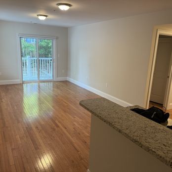 Thomas Interior Painting - A spacious, empty living room features hardwood flooring, a sliding glass door leading to a balcony, and neutral-colored walls. - Painters Near Me - Milton, Massachusetts